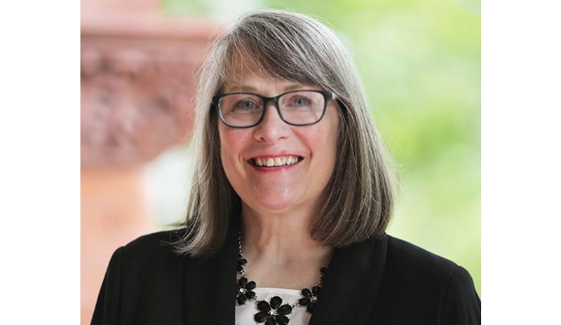 Smiling woman with glasses and shoulder-length gray hair in a black blazer, outdoors, wearing a black flower necklace.