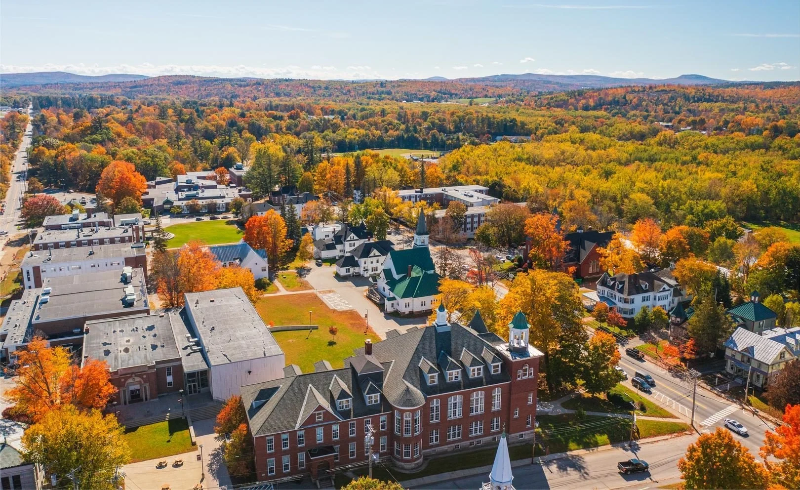Aerial view of a small town campus in fall, with red-brick buildings, white church steeples, and vivid orange and yellow trees across the landscape.