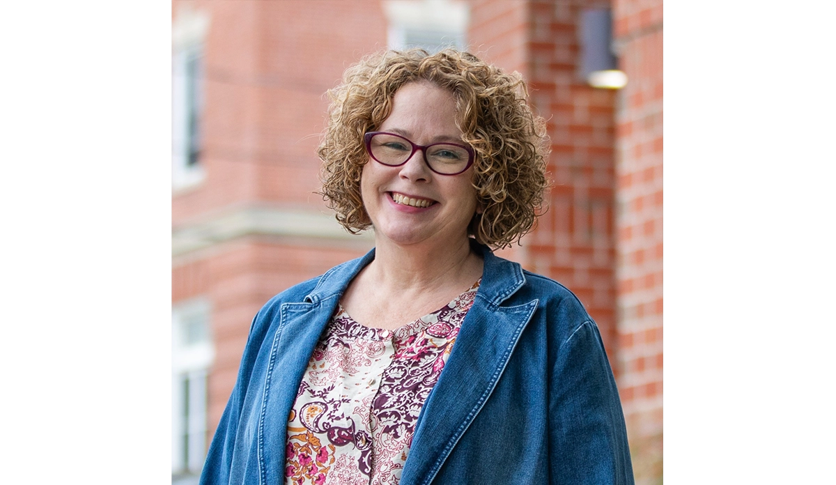 Smiling woman with curly hair and purple glasses wearing a denim jacket outdoors, brick building in the background.