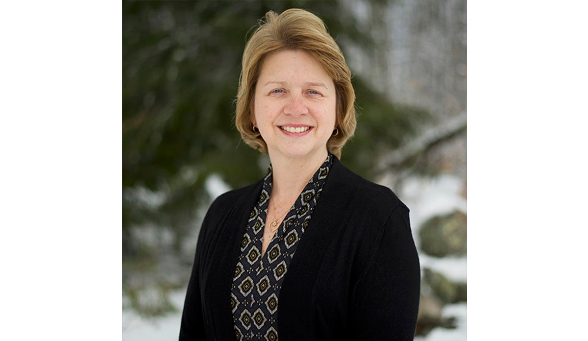Smiling woman in a black blazer and geometric patterned blouse, posing outdoors in a wintery, snowy setting.