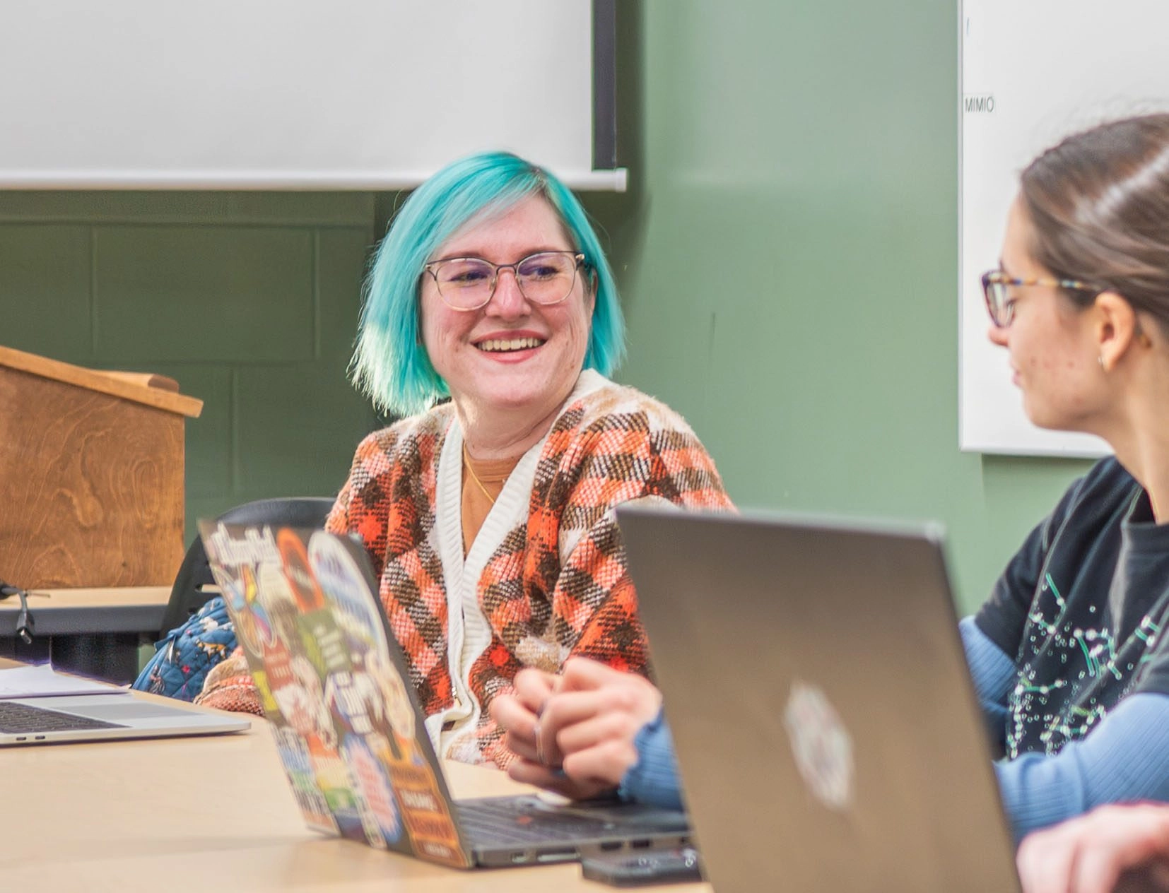 Smiling woman with turquoise hair and glasses sits at a table with laptops and a colorful brochure, talking with a colleague in a classroom setting.
