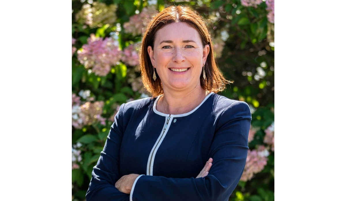 Professional woman with arms crossed, smiling outdoors with pink flowers in the background outside.