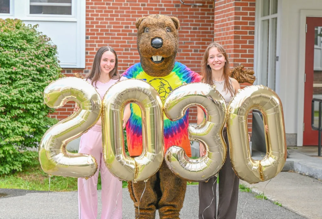 Two smiling young women pose with a Farmington beaver mascot holding large gold balloon numbers saying 2030 outdoors in front of a brick building.