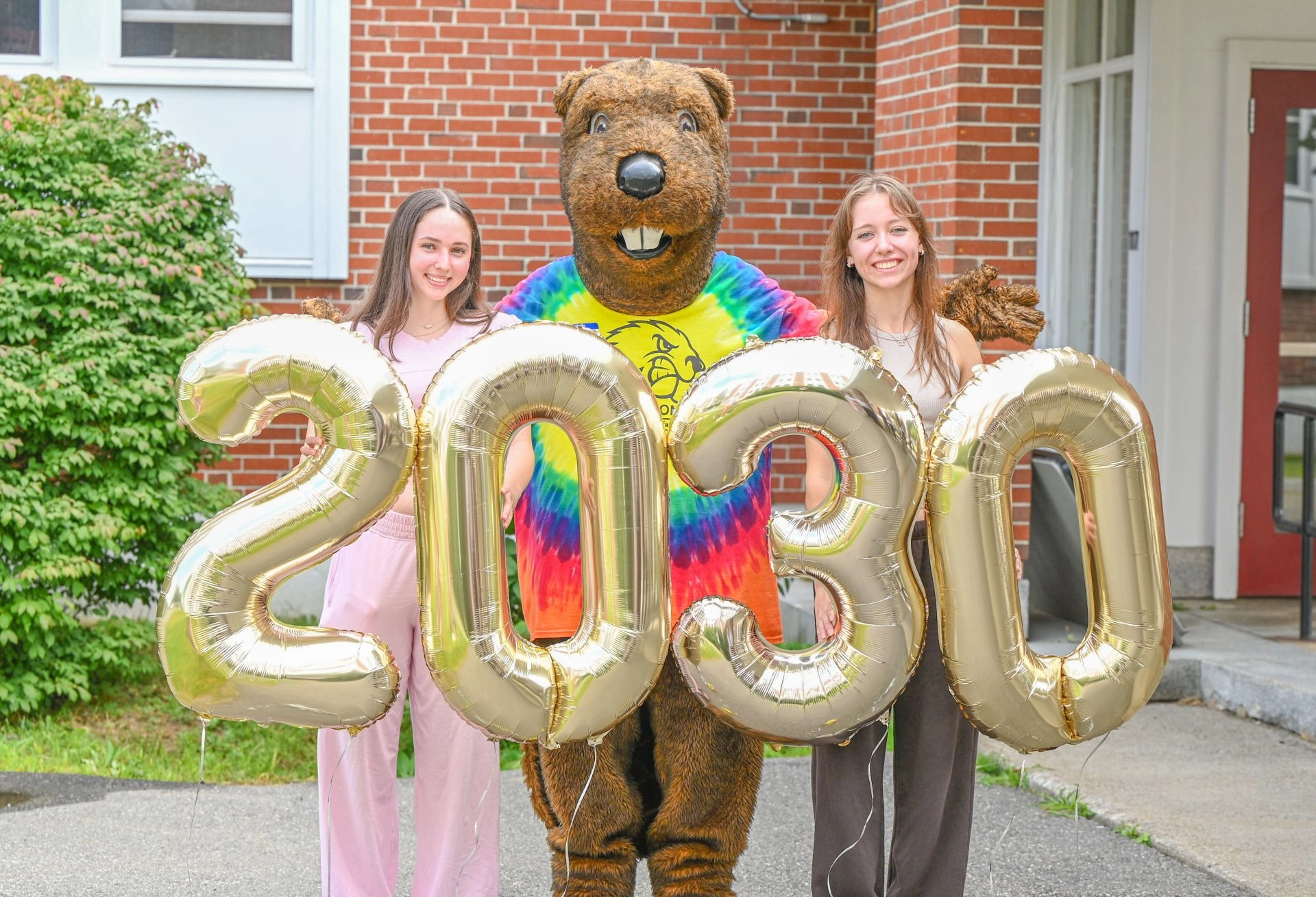 Two smiling young women pose with a Farmington beaver mascot holding large gold balloon numbers saying 2030 outdoors in front of a brick building.