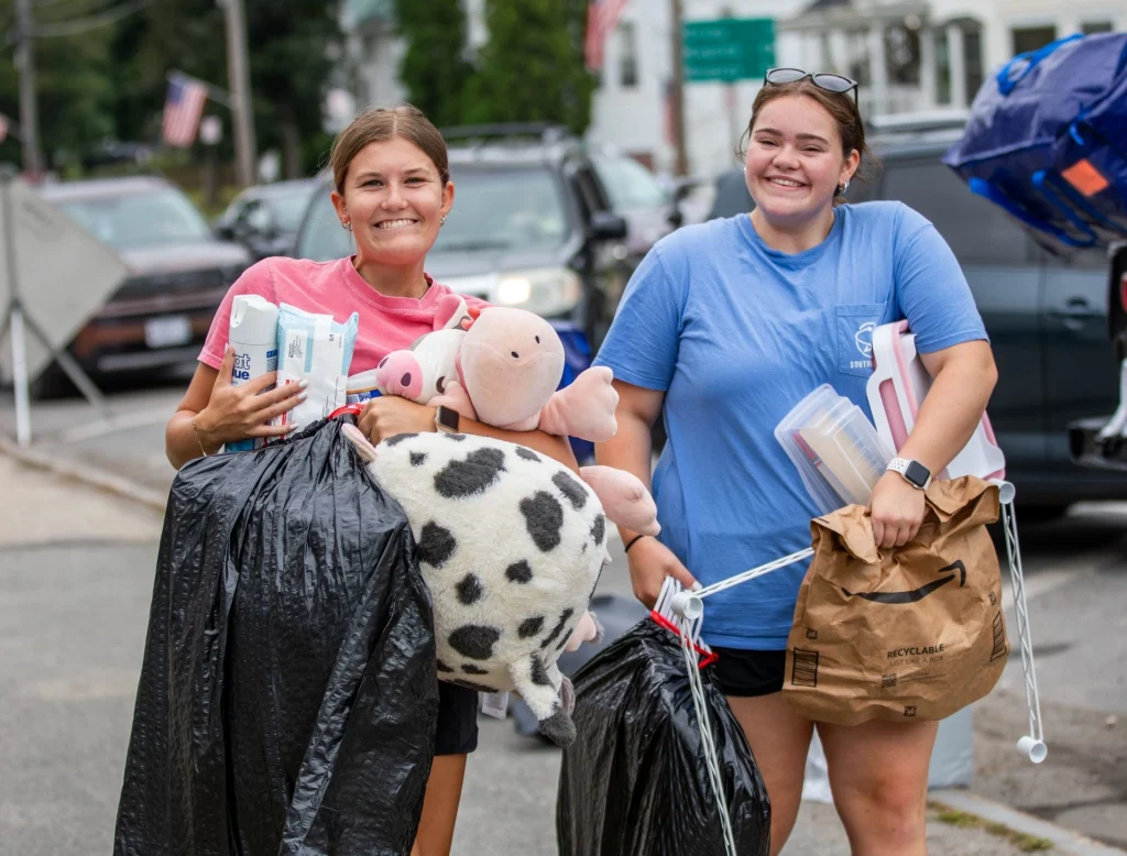 Two smiling students carrrying their belongings to their new residence hall on campus.