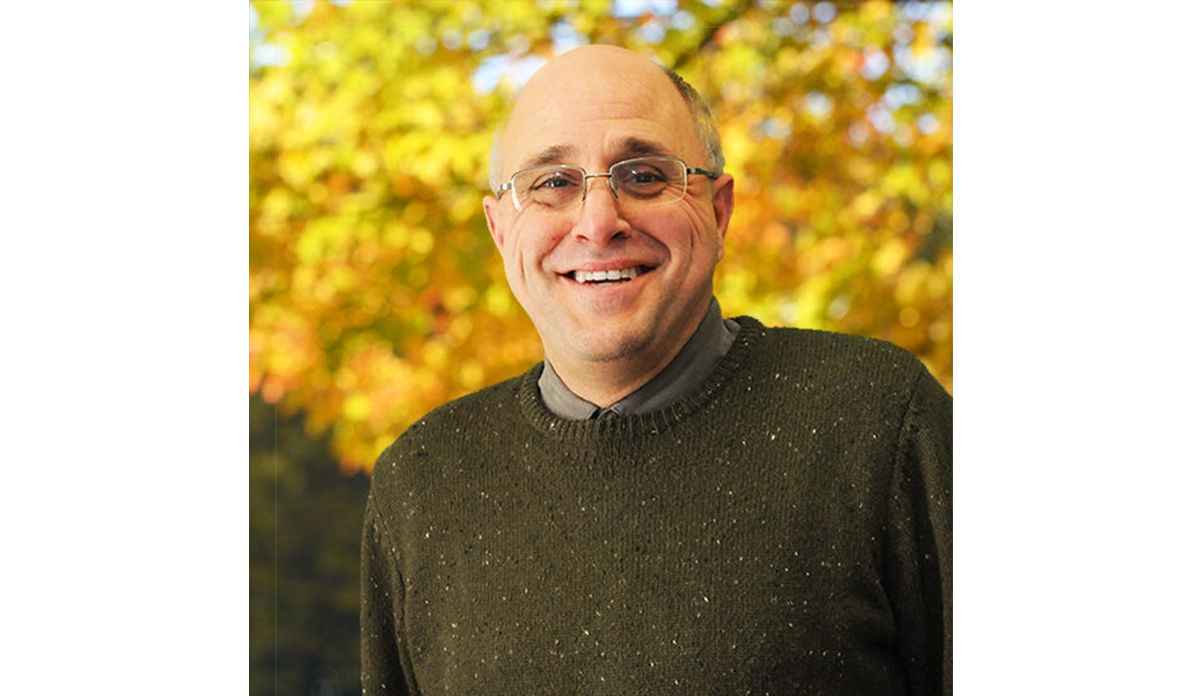 Smiling man wearing glasses outdoors with autumn foliage in the background