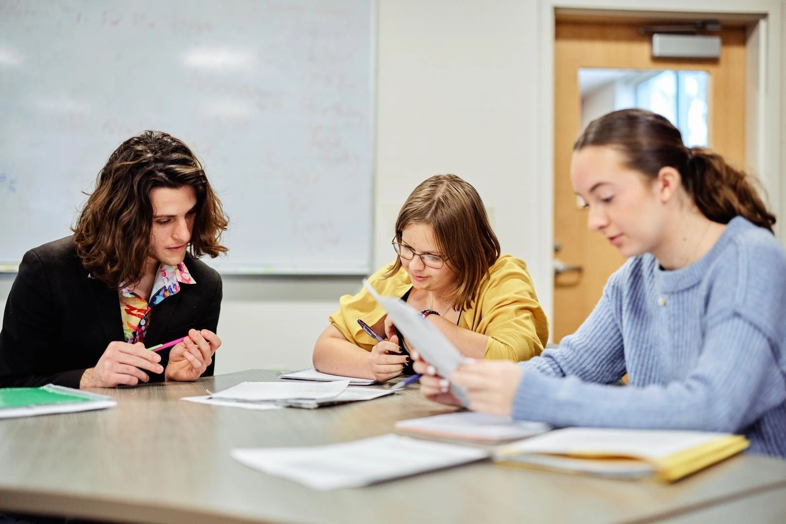 Three students sit at a table, reviewing papers and taking notes in a classroom setting.