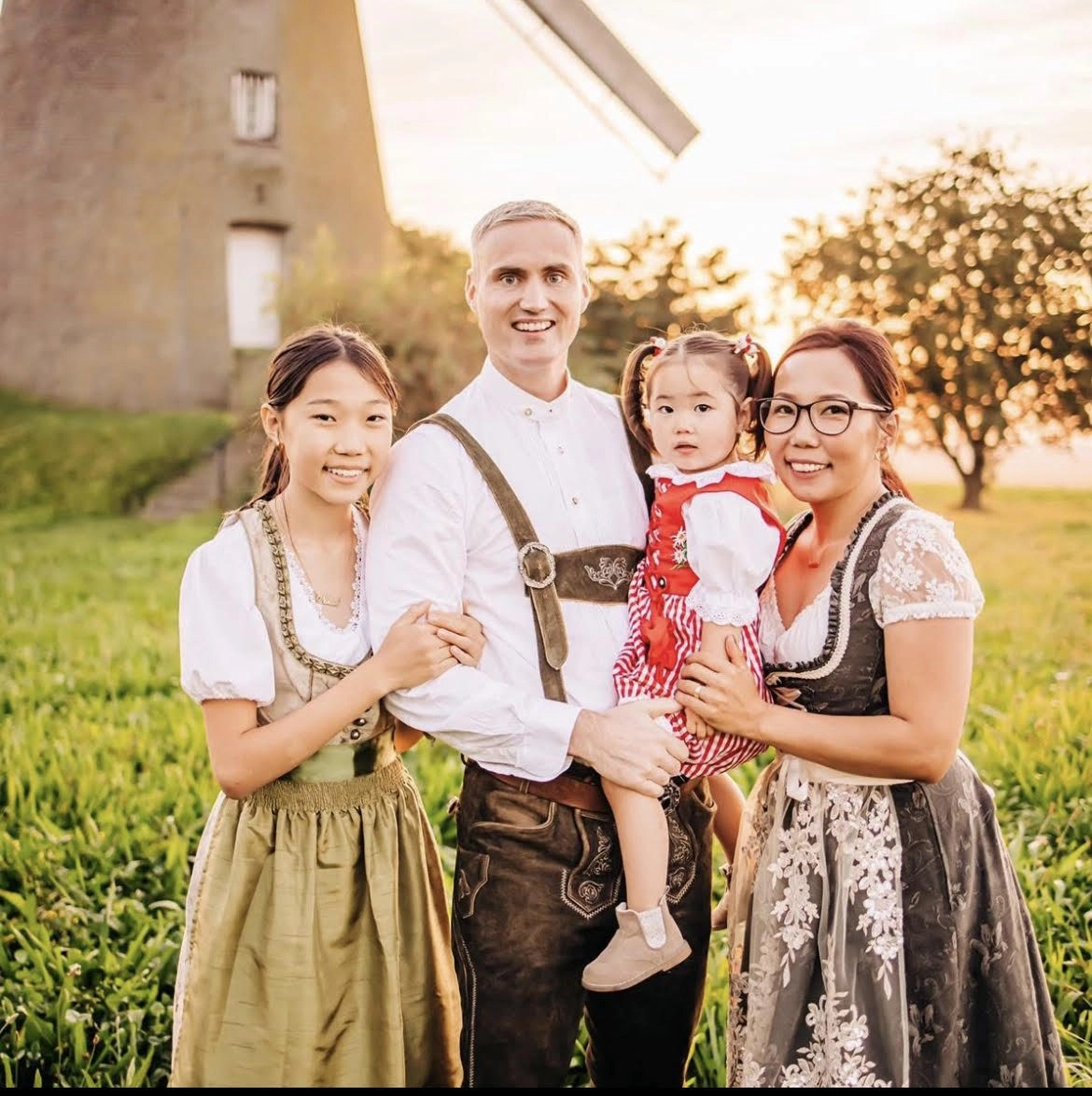 Family of four in traditional Bavarian dress (dirndls and lederhosen) posing in a sunlit field with a windmill in the background, holding a young girl.
