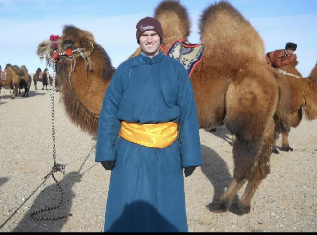 Man in a blue traditional robe with an orange sash posing in a desert with camels in the background