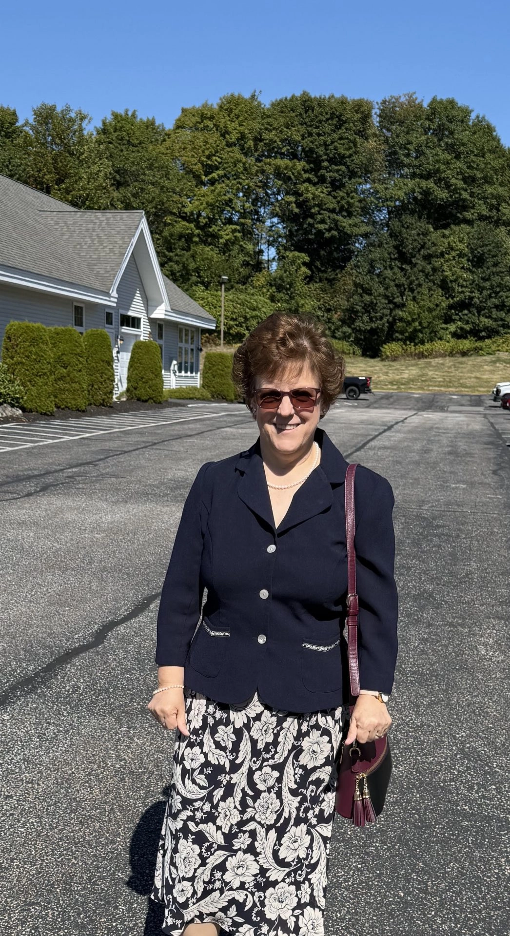 Woman in a navy blazer and floral skirt standing in a parking lot on a sunny day, smiling at the camera with sunglasses and a burgundy handbag.