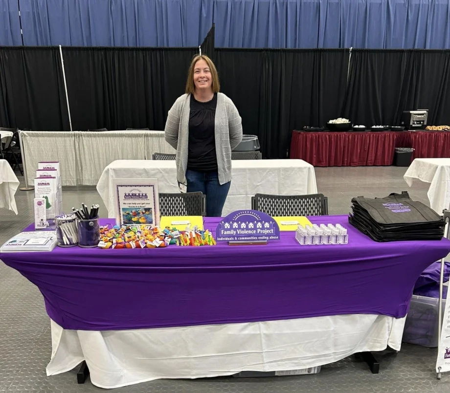 Brunette woman with long bob standing behind table with purple table cloth and Family Violence Project table display