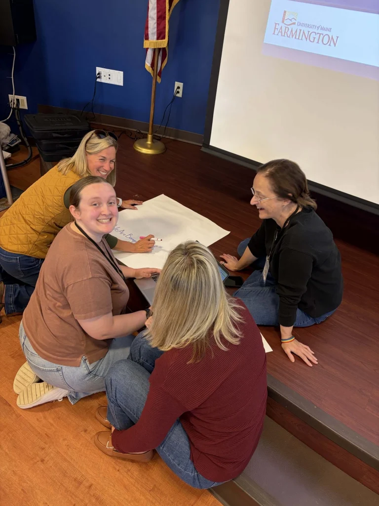 People seated on the floor around a large white poster, writing with markers, while a projector screen showing University of Maine Farmington is in the background.