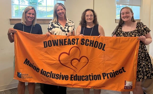 Four women stand indoors, smiling, while holding an orange banner that reads Downeast School Maine Inclusive Education Project,