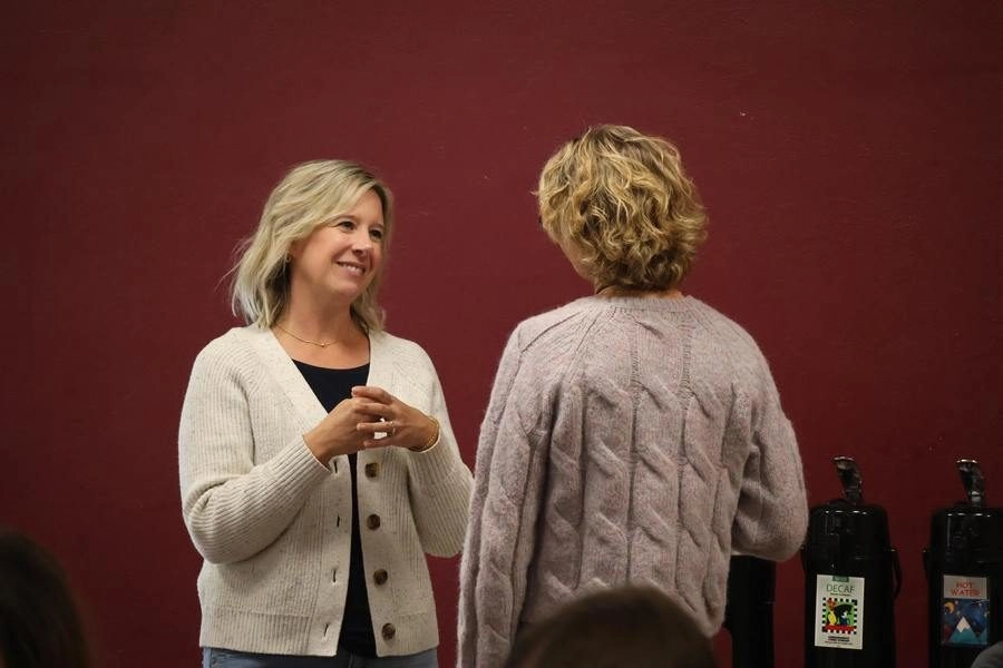 Two women converse in a casual setting, one facing forward with a light cardigan while the other has back to camera against a red wall.