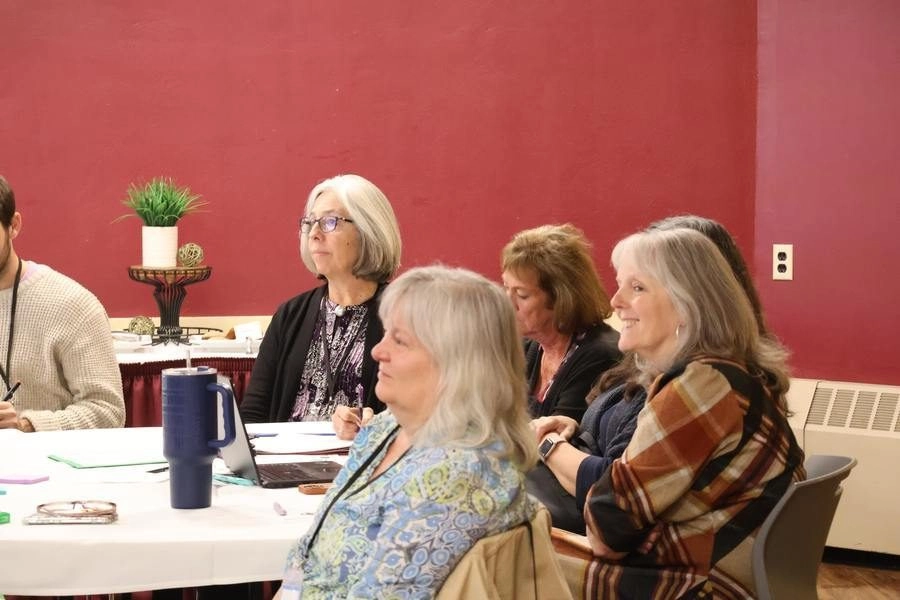 Group of adults seated around a round table in a meeting room, engaging in a workshop or discussion with papers and a laptop visible.