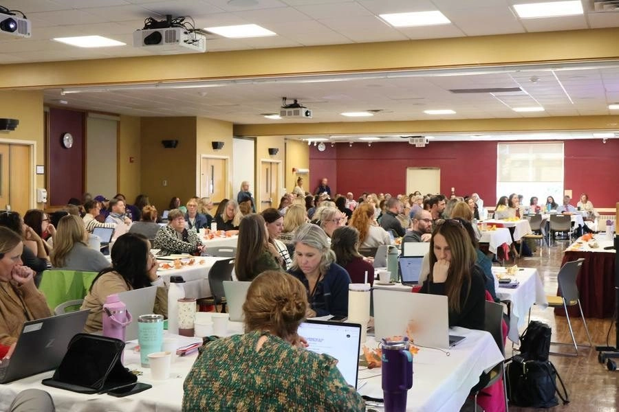 Large room full of attendees seated at round tables with laptops and cups during a conference or workshop.
