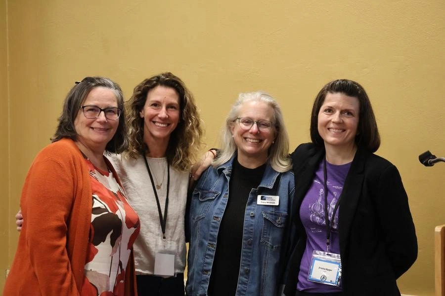 Four women stand together, smiling for a photo in a beige-walled room.