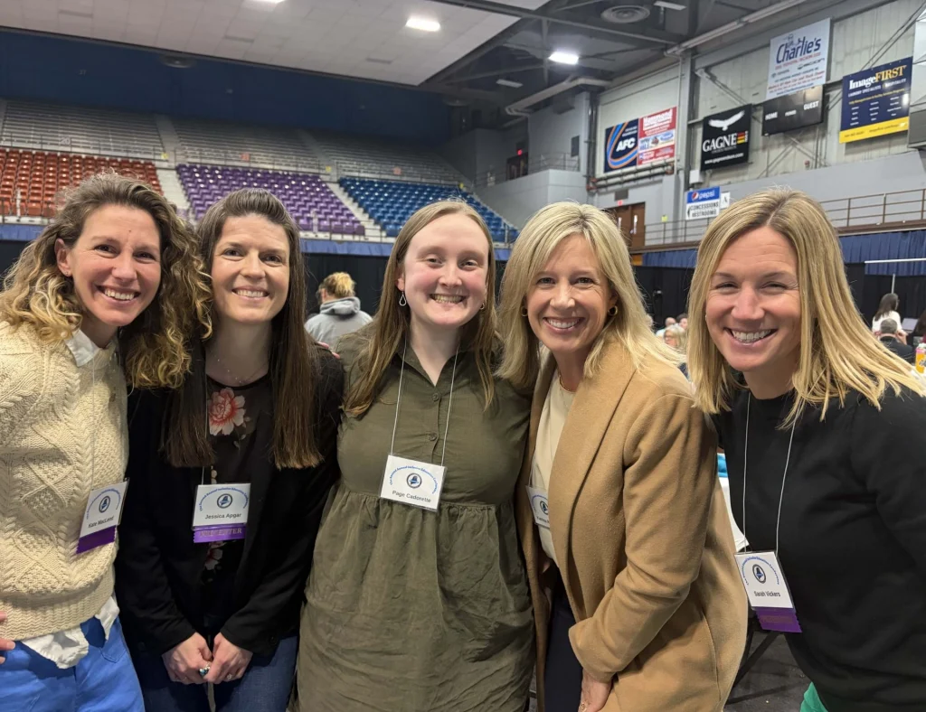 Five smiling women wearing name badges posing for a group photo at an indoor event, with arena seating and banners in the background.