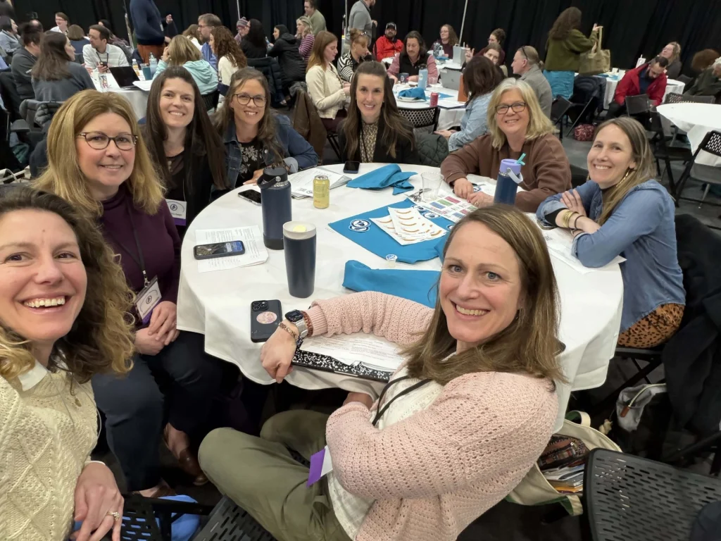Group of smiling adults around a round table at a conference or event, taking a selfie together; name badges, blue napkins, canned drinks and papers visible on the table.