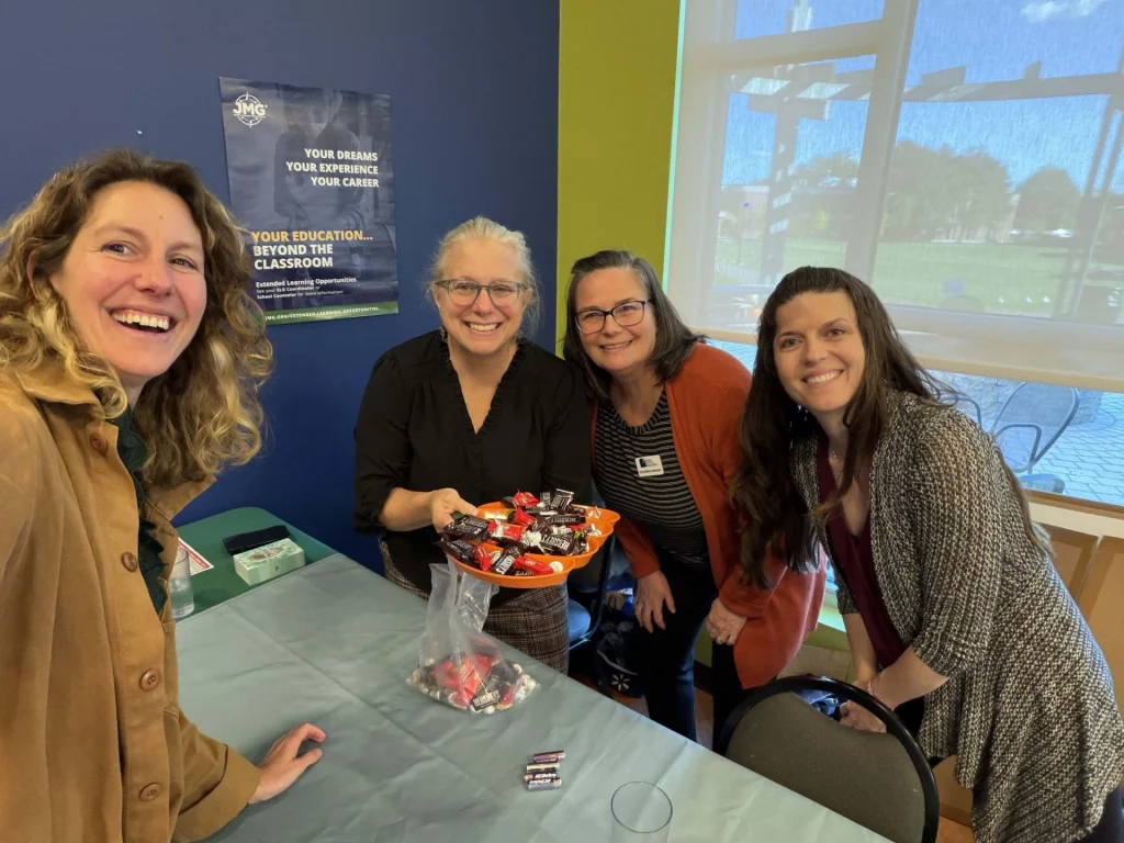 Four women smile at a table with a bowl of wrapped candies in a bright room with blue walls and a poster behind them.
