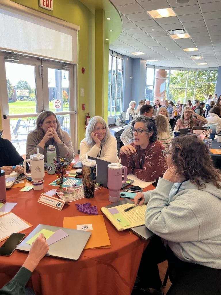 Crowded room of adults seated at orange tables during a workshop, with large windows and greenery outside.