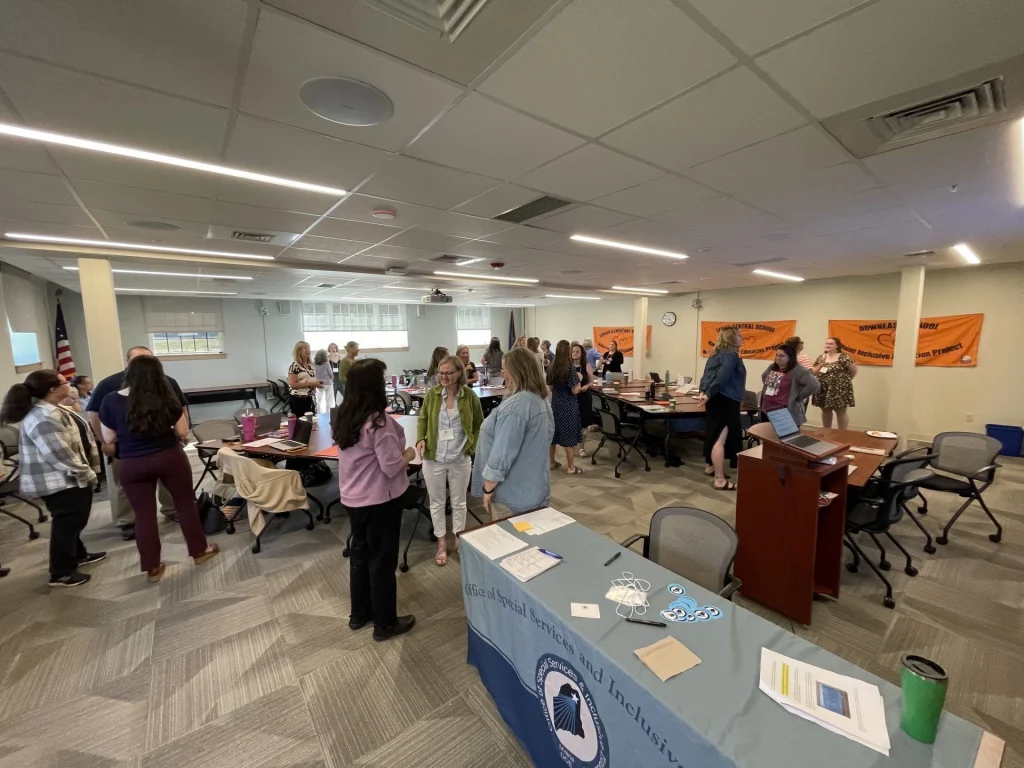 Group of adults mingling in a bright conference room; orange banners on the wall and an American flag in the corner nearby.
