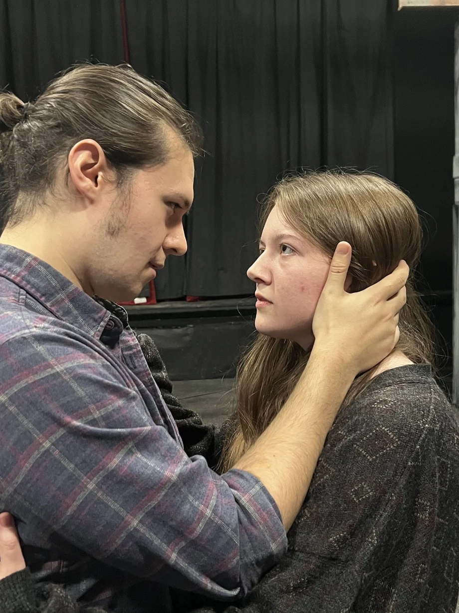 Man holding a young woman's face in his hands, they look into each other's eyes in a dark backstage setting.