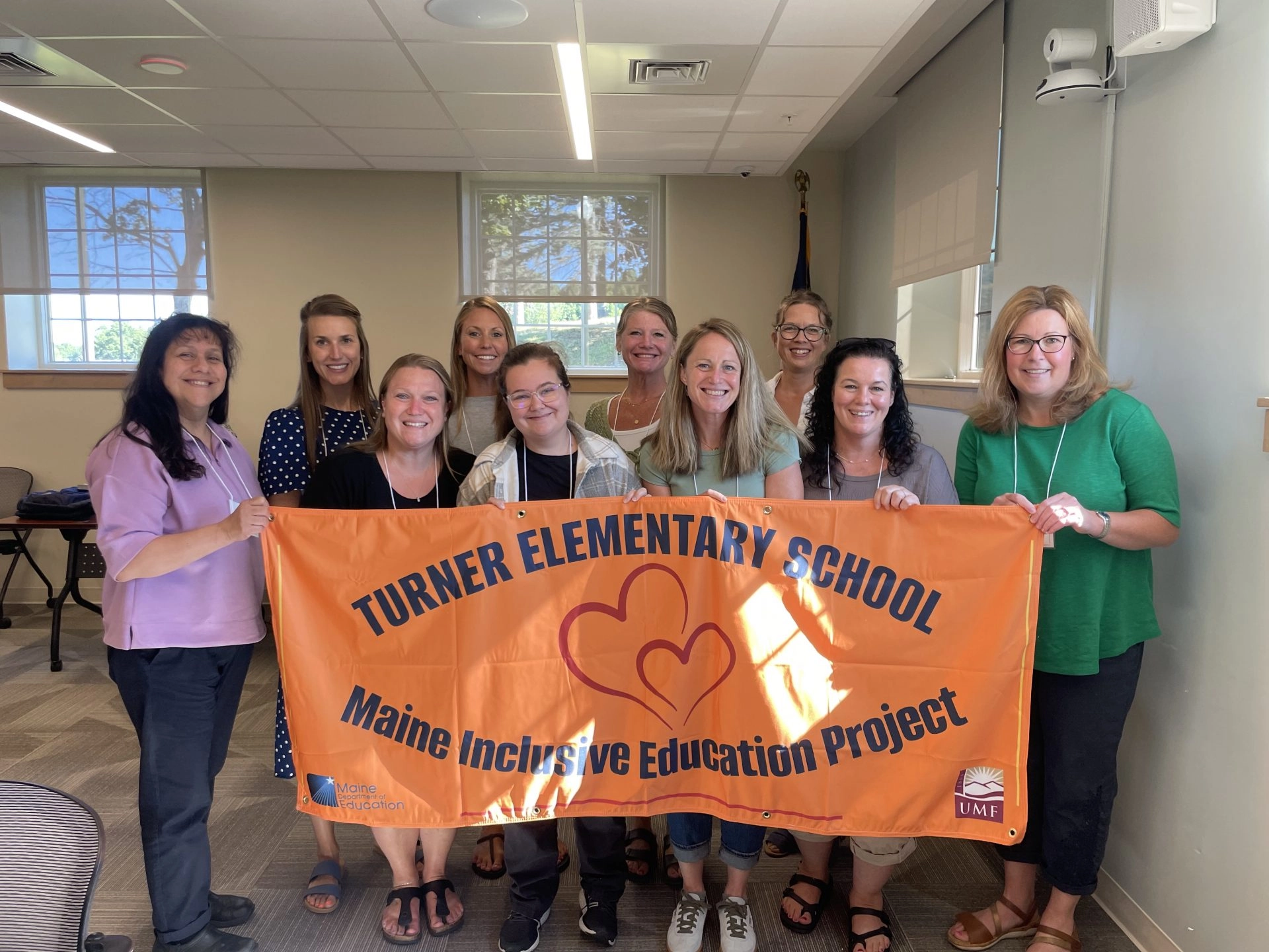 Group of ten adults posing indoors with an orange banner reading Turner Elementary School and Maine Inclusive Education Project.