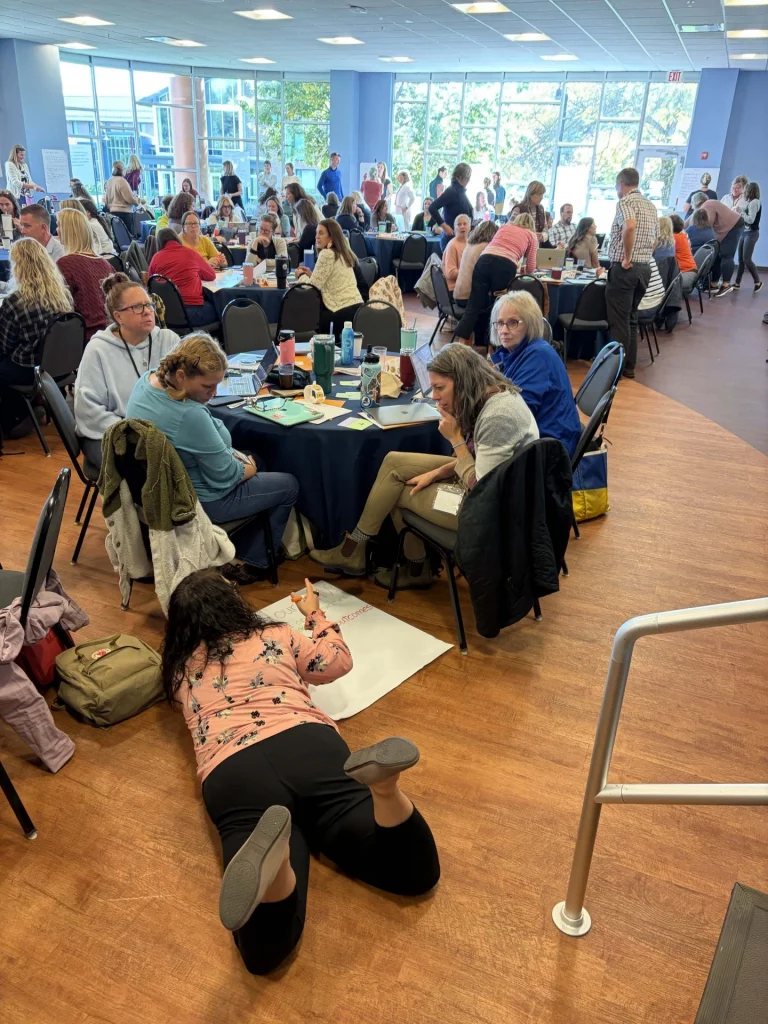 A crowded workshop space with many people seated at round tables, chatting and working on laptops and papers.
